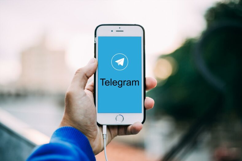 Foto: Nahaufnahme eines iPhone in Weiß, das eine Person in der Hand hält, mit dem Startbildschirm der Telegram-App.